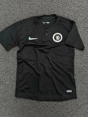 Svart Chelsea FC träningströja Nike - Snygg svart träningströja från Nike med Chelsea FC-logga på bröstet och knappslå vid halsen. Tröjan har korta ärmar och är tillverkad i ett lätt, ventilerande material som passar perfekt till fotbollsträning eller gymmet.