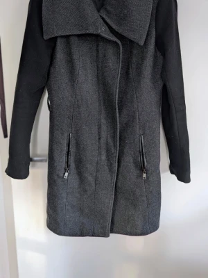 Svart och grå kappa med bred krage - Stilren kappa i svart och grått med bred, asymmetrisk krage och två dragkedjeförsedda fickor framtill. Kappan har raka ärmar och en modern, något figursydd passform. Perfekt för dig som gillar minimalistisk design och vill ha en snygg jacka till hösten. Väldigt fint skick 