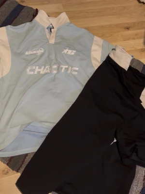 Ljusblå Chaotic t-shirt med tryck - Säljer en ljusblå t-shirt från Chaotic med vita detaljer och tryck på bröstet. T-shirten har korta ärmar, vit krage och sportig design. Perfekt för dig som gillar streetwear eller sportig stil.