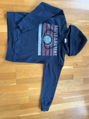 Mörkblå Saint Laurent hoodie - Snygg mörkblå hoodie från Saint Laurent med tryck framtill i rött, vitt och grått. Klassisk känguruficka och huva. Perfekt för en avslappnad streetstyle-look. Mjukt material och långa ärmar.