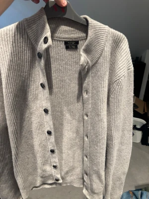 Kofta från Massimo Dutti - Ljusgrå ribbstickad kofta från Massimo Dutti med svarta knappar framtill. Materialet är mjukt och skönt, och modellen är klassisk och enkel att matcha. Använd väldigt väldigt få gånger. Storlek L men skulle nog säga passar bättre M