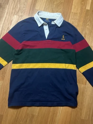 Randig långärmad topp Polo Ralph Lauren - Säljer en långärmad topp från Polo Ralph Lauren med breda ränder i marinblått, rött, grönt och gult. Klassisk vit krage och broderad logga på bröstet. Perfekt för dig som gillar preppy stil och vill sticka ut med färg.