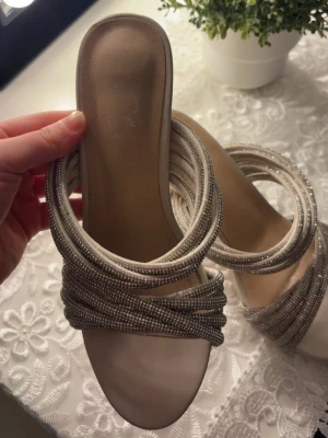 Beige sandaler med glittriga remmar - Säljer ett par snygga beige sandaler med flera glittriga remmar i silver. Sandalerna har öppen tå och låg klack, vilket ger en trendig look. Materialet är en mix av textil och syntet, och remmarna är dekorerade med små pärlor för extra bling.