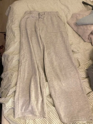 Grå mjukisbyxor med raka ben - Säljer ett par grå mjukisbyxor med raka ben och elastisk midja med dragsko. Byxorna har stora fickor framtill och är gjorda i ett mjukt och bekvämt material, perfekt för chill dagar. Passar dig som gillar en avslappnad och enkel stil.