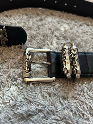 Chrome bälte - Coolt svart bälte från Chrome Hearts med unika silvriga detaljer och spänne. Bältet har små svärdformade dekorationer längs hela remmen och är gjort i skinn. Perfekt accessoar för att lyfta din outfit.