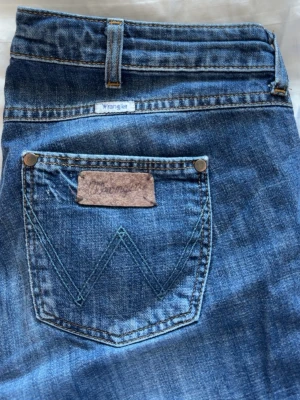 Blå jeans från Wrangler - Ett par klassiska blå jeans från Wrangler med fem fickor och tydliga kontrastsömmar. Jeansen har en snygg slitning på vänster lår och ikonisk W-detalj på bakfickan. Tillverkade i slitstarkt denimtyg som ger en cool och avslappnad look.