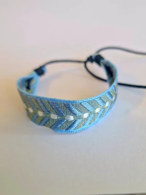 Blått tygarmband med mönster - Snyggt armband i tyg med ljusblå och gråa toner samt vita detaljer. Armbandet har ett chevronmönster och justerbar knytning baktill för perfekt passform. Perfekt accessoar för en avslappnad och trendig stil.