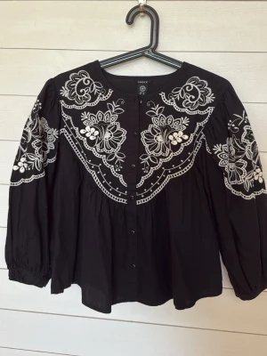 Svart broderad blus från Lindex - Svart blus från Lindex med vita broderier i blommigt mönster över axlar, bröst och ärmar. Blusen har rund halsringning, knäppning framtill och långa puffärmar. Tillverkad i mjuk bomull och har en avslappnad passform.