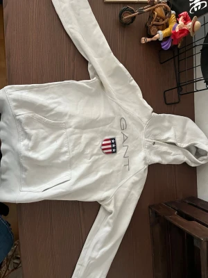 Vit hoodie från GANT med logga - Snygg vit hoodie från GANT med broderad logga och amerikanskt inspirerat emblem på bröstet. Klassisk känguruficka framtill och huva med dragsko. Perfekt för en avslappnad och stilren look.