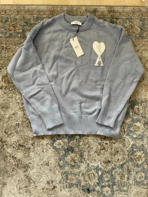 Ami Paris tröja - Snygg ljusblå stickad tröja från Ami med rund halsringning och ribbade muddar. På bröstet finns ett vitt hjärta och bokstaven A som broderad detalj. Tröjan är i mjukt material och har en avslappnad passform. Perfekt för dig som gillar stilrena plagg med en twist.