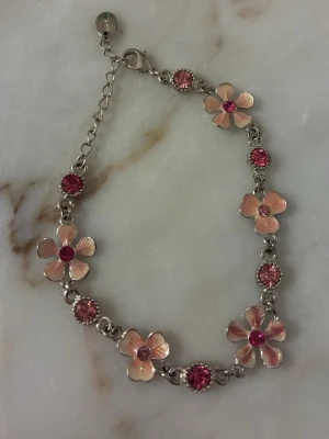 Silver armband med rosa blom stenar - Sött armband med silverfärgad kedja, dekorerat med rosa blommor och gnistrande rosa stenar. Blommorna har en metallisk finish och armbandet har justerbar längd med karbinlås. Perfekt för dig som gillar en feminin och lekfull stil.