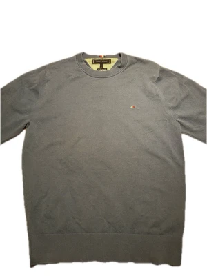 Grå tröja från Tommy Hilfiger - Klassisk grå tröja från Tommy Hilfiger med rund halsringning och diskret logga broderad på bröstet. Tröjan har långa ärmar och ribbade muddar vid ärmslut och nederkant. Perfekt för en clean och stilren look.