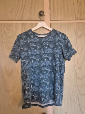 Blå mönstrad t-shirt med palmblad - Snygg blå t-shirt med vitt palmbladsmönster. Perfekt för dig som gillar en avslappnad och trendig stil. T-shirten har rund hals och korta ärmar. Skön och lätt att bära till jeans eller shorts.     Alla kläder rollas innan dom skickas hem då det finns katt i hemmet🐈 Jag stryker och tvättar INTE innan jag skickar det får du stå för själv så det blir som du vill ha det🤗
