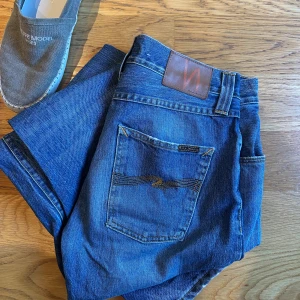 Blå nudie jeans - Säljer ett par klassiska blå jeans från Nudie Jeans med raka ben och femficksmodell. Jeansen har en snygg tvätt och kontrastsömmar samt Nudie-logga på bakfickan och läderpatch i midjan. Perfekta för en avslappnad och stilren look.