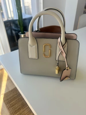 Grå handväska från Marc Jacobs - Snygg handväska från Marc Jacobs i ljusgrått läder med guldfärgade detaljer och ett dekorativt hänglås. Väskan har två handtag, rosa insida och en avtagbar tag. Perfekt för dig som vill ha en stilren och trendig accessoar.