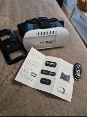 VR BOX VR-glasögon med kontroll - VR-glasögon från VR BOX i vit och svart plast med justerbara band. Medföljer en svart handkontroll och instruktioner. Perfekt för att uppleva spel och filmer i 3D med mobilen. Levereras i originalkartong.