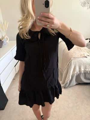 Svart kort klänning med volang - Säljer en svart kort klänning med volangkant nedtill och knyt i halsen. Klänningen har korta ärmar och en lös passform som ger en avslappnad look. Perfekt för dig som gillar enkel men stilren design.