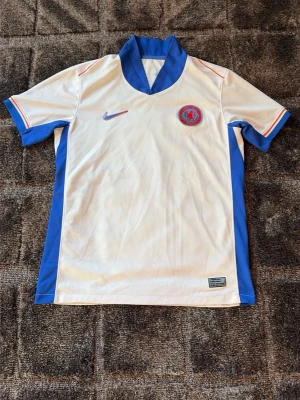 Chelsea bortatröja João Félix 14 - Chelsea FC bortatröja från Nike med João Félix och nummer 14 på ryggen. Tröjan är vit med blå detaljer, blå krage och röda inslag på ärmarna. Tillverkad i lätt och ventilerande material, perfekt för fotboll eller träning.har en defekt på ryggen där numret står så har siffrorna börjat flagnat lite där av priset!