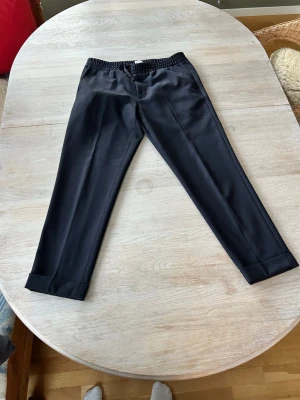 Filips k - Svarta kostymbyxor med resår - Stilrena svarta kostymbyxor med resår i midjan och raka ben. Byxorna har pressveck framtill och uppvikta benslut för en modern look. Perfekta för dig som vill ha en dressad men avslappnad stil.