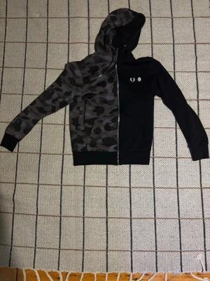 Svart och camo hoodie Fred Perry x BAPE - Unik hoodie med halvsvart och halvcamouflage design från Fred Perry x A Bathing Ape. Dragkedja framtill, två fickor och huva. Materialet är mjukt och bekvämt, perfekt för att sticka ut. Logotyp på bröstet och text på ryggen.