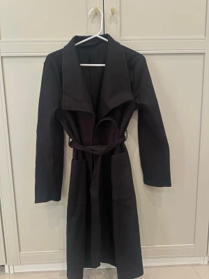 Svart lång kappa med knytskärp - Stilren svart lång kappa med bred krage och knytskärp i midjan. Kappan har en rak passform och är tillverkad i ett mjukt tyg som ger en snygg siluett. 