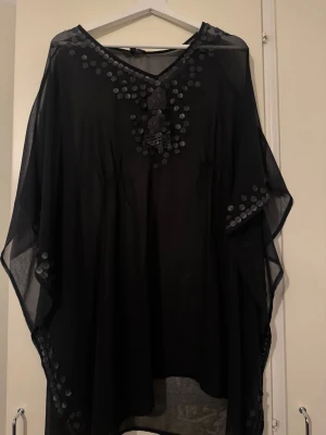 Svart strandponcho med paljetter - Snygg svart strandponcho i transparent, lätt material med dekorativa svarta paljetter runt halsringningen och längs kanterna. Ponchon har vid passform och v-ringning, perfekt att slänga över bikinin på stranden.