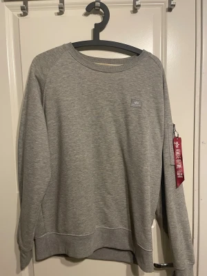 Grå sweatshirt från Alpha Industries - Snygg grå sweatshirt från Alpha Industries med rund halsringning och liten logotyp på bröstet. Tröjan har en klassisk passform, ribbade muddar och en ikonisk röd tag på ärmen. Perfekt för en avslappnad streetwear-look.