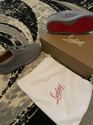 Gråa Louboutin sneakers med nitar - Snygga gråa sneakers från Christian Louboutin med klassisk röd sula och coola silverfärgade nitar på tån. Skorna är i mocka och har en modern, stilren design som sticker ut. Perfekta för dig som vill ha något unikt och trendigt i garderoben. Säljer för använder it endast provat och har ej kläder som matchar med skorna.