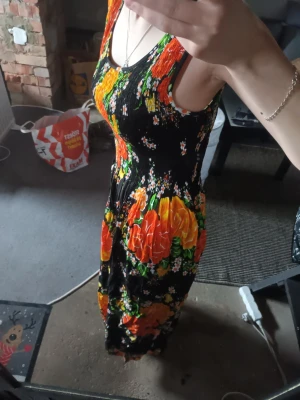 Svart långklänning med blommor - Färgstark långklänning med orange, gula och gröna blommor på svart botten. Ärmlös modell med rund halsringning och figurnära överdel. Storlek vet jag ej, men passar för S och M. Perfekt för dig som vill ha en somrig look inför sommaren🌷