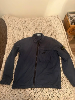 Blå overshirt från Stone Island - Snygg blå overshirt från Stone Island med dragkedja framtill och klassisk krage. Bröstficka med knapp och ikonisk logotyp-patch på vänster ärm. Perfekt lager för en streetwear-look.