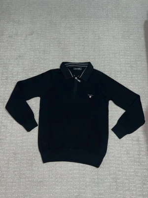 Svart ziptröja med krage från GANT - Snygg svart tröja från GANT med halv dragkedja och klassisk krage. Tröjan har vita ränder på kragen och GANT-logga broderad på bröstet. Perfekt för en stilren och sportig look.