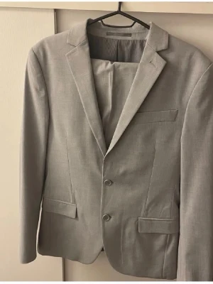 Ljusgrå kostym från dressman  - Ljusgrå kavaj med två knappar framtill, smala klassiska slag och tre fickor. Kavajen har en stilren och modern design, perfekt för dig som gillar en clean look. Materialet är mjukt och ger en bekväm känsla hela dagen.