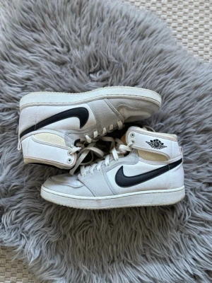 Nike Air Jordan 1 vita och svarta - Säljer ett par klassiska Nike Air Jordan 1 sneakers i vitt med svarta detaljer och ikonisk swoosh på sidan. Skorna har högt skaft, snörning och är tillverkade i en mix av läder och textil. Perfekta för dig som gillar streetwear och tidlös design.