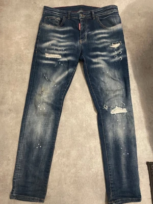 Blå jeans från Dsquared2 - Säljer ett par blå jeans från Dsquared2 med slitningar, hål och färgstänk för en riktigt edgy look. Jeansen har fem fickor, låg midja och smal passform. Snygga röda detaljer vid fickan och klassisk knappgylf. Perfekta för dig som gillar streetstyle.