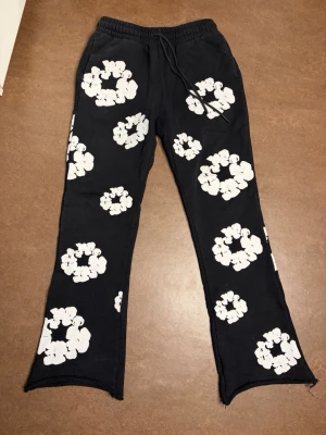 Svarta mjukisbyxor Denim Tears - Unika svarta mjukisbyxor från Denim Tears x Readymade med stora vita blommönster över hela byxorna. Byxorna har resår i midjan med dragsko och raka ben med råa kanter nertill. Perfekta för dig som vill sticka ut med en cool streetwear-look.