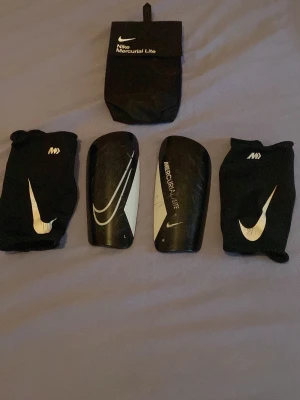 Nike Mercurial Lite benskydd svart - Säljer ett par svarta Nike Mercurial Lite benskydd med vita detaljer och tillhörande svarta sleeves. Benskydden har en lätt och smidig form med stötdämpande baksida och kommer med en praktisk svart förvaringspåse. Perfekt för fotbollsträning eller match.