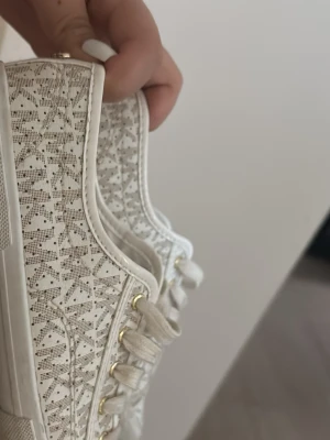 Vita spets-sneakers från Michael Kors - Snygga vita sneakers från Michael Kors med detaljer i spets och guldfärgade nitar baktill. Skorna har en låg profil och klassisk gummisula. Perfekta för dig som vill ha en unik touch på dina sneakers.