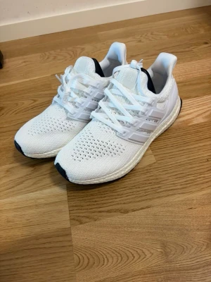 Vita Adidas Ultraboost sneakers - Snygga vita Adidas Ultraboost sneakers med stickad ovandel i mesh för extra andningsförmåga. Skorna har klassiska tre ränder på sidan, vit sula med Boost-dämpning och svarta detaljer på yttersulan. Perfekta för dig som gillar sportig och clean stil.