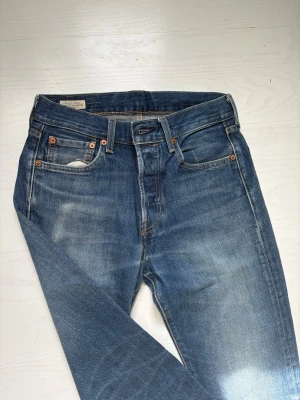 Blå raka jeans från Levi's - Klassiska blå jeans från Levi's med raka ben och fem fickor. Jeansen har en snygg tvättad look med slitningar och kopparfärgade nitar. Tillverkade i slitstark denim med normal midja och gylf med knappstängning. Storlek. W29 L32