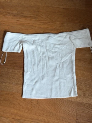 Vit offshoulder topp från H&M Divided - Snygg vit offshoulder topp från H&M Divided i ribbad bomull. Toppen har korta ärmar och sitter tight över axlarna, perfekt för en trendig look. Enkel att matcha med både jeans och kjol.