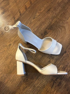 Creamvita pumps med blockklack och rem - Stilrena creamvita pumps från NA-KD med bred blockklack och fyrkantig tå. Skorna har en enkel rem över tårna och justerbar ankelrem med spänne. Tillverkade i slätt konstläder som ger en modern look. Helt oanvända