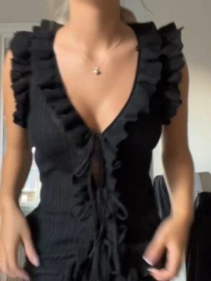 Svart ribbad topp med volang  - Säljer en svart ribbad topp från Zara med volangdetaljer längs v-ringningen och axlarna. Toppen har snörning framtill och är ärmlös.