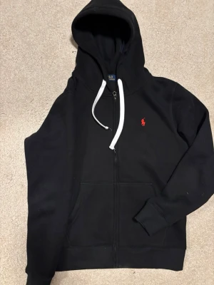 Svart Polo Ralph Lauren zip - Svart hoodie med dragkedja från Polo Ralph Lauren. Klassisk design med vit dragsko i huvan och röd broderad logga på bröstet. Tillverkad i mjukt bomullsmaterial och har känguruficka framtill. Perfekt för en chill och stilren look.