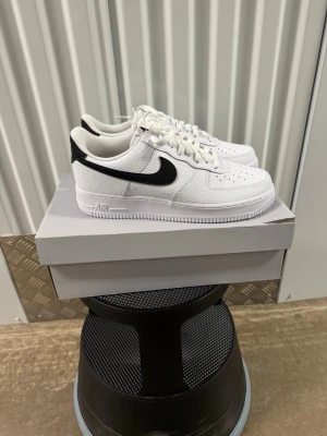 Nike Air Force 1 vita med svart swoosh - Klassiska Nike Air Force 1 sneakers i vitt läder med svart swoosh på sidan. Lågt skaft, perforerad tåbox för ventilation och vit sula. Tidlös design som passar till det mesta och ger en clean look.
