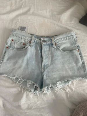 Ljusblå Levi's jeansshorts med fransar - Säljer ett par ljusblå jeansshorts från Levi's med hög midja och råa fransade kanter. Klassisk femficksmodell med knapp och dragkedja framtill samt Levi's patch bak. Perfekta för sommaren!