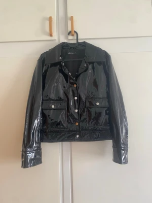 Svart glansig skinnjacka från ginatricot - Snygg svart skinnjacka med glansig finish och klassisk krage. Jackan har två stora fickor framtill med lock och silverfärgade knappar. Rak passform och långa ärmar ger en cool och modern look.