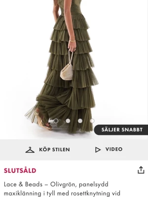 Olivgrön maxiklänning från Lace & Beads - Olivgrön långklänning i tyll från Lace & Beads med flera lager volanger och breda axelband som kan knytas i stora rosetter. Klänningen har en figurnära överdel korset och ett svepande, romantiskt fall. Perfekt för dig som vill sticka ut med en unik och elegant look. Perfekt till fest eller bal.