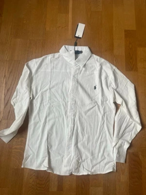 Vit skjorta från Polo Ralph Lauren - Klassisk vit skjorta från Polo Ralph Lauren med broderad marinblå logga på bröstet. Skjortan har lång ärm, knappar framtill och är tillverkad i bomull. Perfekt till både jeans och chinos för en clean och stilren look.