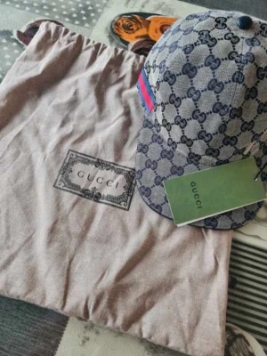 Gucci keps med monogram och röd-blå rand - Säljer en snygg keps från med klassiskt monogram i blått på grå bakgrund. Kepsen har en röd-blå rand på sidan och kommer med originalpåse och tagg. Perfekt för dig som vill ha en exklusiv och trendig accessoar.