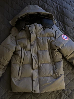 Grå pufferjacka från Canada Goose - Snygg grå pufferjacka från Canada Goose med klassisk logga på ärmen. Jackan har huva, dragkedja och knappar framtill samt två stora fickor. Perfekt för kalla dagar med sin vadderade design och stilrena look.